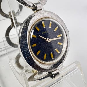 Könnte beinhalten: Eine silberfarbene Armbanduhr mit einem runden, texturierten Gehäuse und einem dunkelblauen Zifferblatt. Das Zifferblatt zeigt das Wort Para, goldfarbene Stundenmarkierungen und dünne goldene Zeiger. Sie hat ein Armband aus großen, ineinandergreifenden silberfarbenen Ringen.