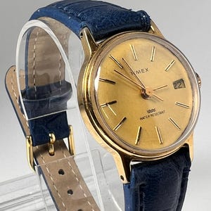 Könnte beinhalten: Eine goldfarbene Timex Armbanduhr mit rundem, goldenem Zifferblatt, goldenen Stundenmarkierungen, einem Datumsfenster auf der Drei-Uhr-Position und einem strukturierten blauen Lederarmband mit goldfarbener Schließe. Das Zifferblatt zeigt Timex und Water Resistant.