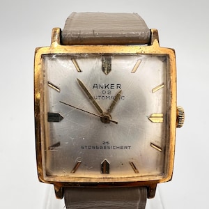 Könnte beinhalten: Eine quadratische, goldfarbene Armbanduhr mit silbernem Zifferblatt, beschriftet mit ANKER 02 AUTOMATIC und 25 STOSSGESICHERT. Die Uhr hat goldfarbene Indizes, schmale Zeiger und ein hellgraues Lederarmband mit sichtbaren Gebrauchsspuren.