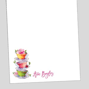 May include: A stack of three teacups with floral designs and a cherry on top. The teacups are stacked on top of each other and are on a white background. The name "Ann Boyles" is written in pink cursive below the teacups.