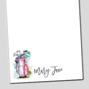 May include: A pink golf bag with golf clubs sticking out of the top, set against a watercolor background of green and blue. The text "Mary Jane" is written in cursive below the bag.