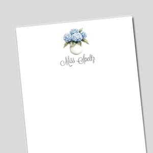 May include: White stationery with a watercolor illustration of blue hydrangeas in a white vase. The text "Miss Speltz" is written in elegant script below the floral arrangement. The stationery is on a white background.