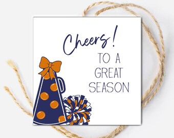 May include: A white square tag with the words "Cheers! To a Great Season" in navy blue script. The tag features a navy blue megaphone with orange accents and a matching pom-pom. A brown string is attached.