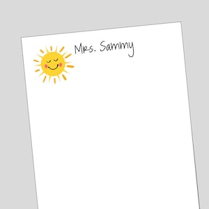 May include: A white notepad with a yellow sun with a smiling face and rosy cheeks. The text "Mrs. Sammy" is written in black ink at the top of the notepad.