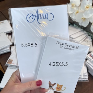 May include: Two white notepad sets with personalized text. The larger notepad is 5.5x8.5 inches and says "Anna". The smaller notepad is 4.25x5.5 inches and says "From the Desk of Dr. Dreskin". The smaller notepad has a cartoon dog and cat on the bottom.