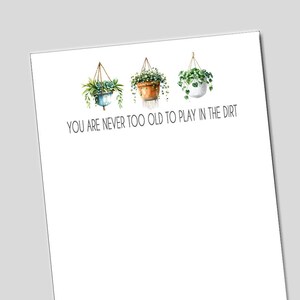 May include: A white print featuring three hanging planters with greenery. The planters are in blue, brown, and white. The text "YOU ARE NEVER TOO OLD TO PLAY IN THE DIRT" is below the planters.