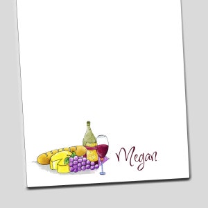 May include: A white notepad with a watercolor illustration of a bottle of wine, a glass of wine, a baguette, a wedge of cheese, and a bunch of grapes. The name "Megan" is written in cursive at the bottom of the notepad.