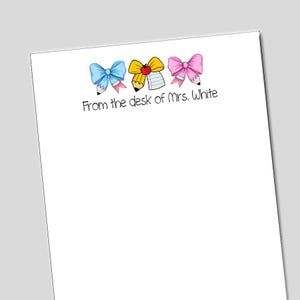 May include: A white sheet of paper with three illustrated pencil bows in blue, yellow, and pink. The text "From the desk of Mrs. White" is written in black cursive below the bows. The bows are detailed with pencil tips and ribbons.