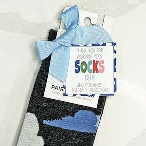 May include: A blue and white gift tag with white polka dots and the text "Thank you for working your socks off!!! And for being toe-tally awesome!" tied to a pair of gray socks with a blue cloud design.