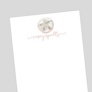 May include: A white card with a pink watercolor sand dollar and the text "casey speltz" in pink cursive.