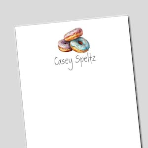 May include: A stack of three donuts with pink, blue, and brown frosting and sprinkles. The donuts are on a white background with the name "Casey Speltz" written in cursive.