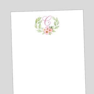 May include: A white notepad with a pink floral wreath and the letter "C" in the center. The wreath is made of green leaves and pink flowers.