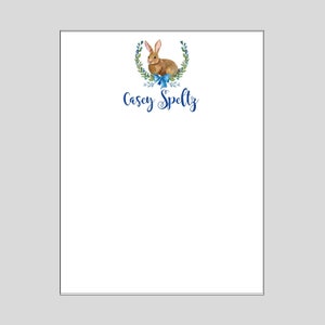 May include: A white notepad with a blue wreath of leaves and berries surrounding a brown rabbit with a blue bow. The name "Casey Speltz" is written in blue cursive lettering below the rabbit.