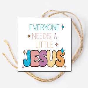 May include: A white card with the text "Everyone needs a little Jesus" in colorful, retro-style lettering. The text is surrounded by small, sparkling stars.