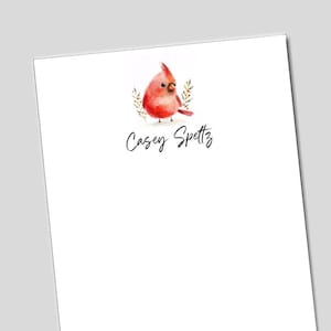 May include: White stationery with a watercolor illustration of a red cardinal bird and greenery. The name "Casey Spettz" is written in cursive below the bird. The design is simple and elegant.