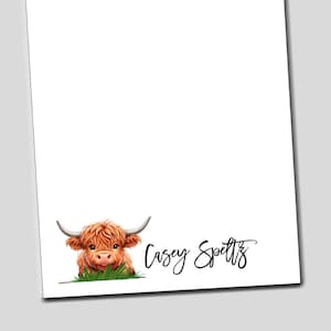 May include: A white notepad with a cute cartoon Highland cow peeking out from the bottom. The notepad has the name "Casey Speltz" written in cursive at the bottom.