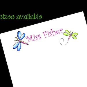 May include: A white card with the text "Miss Fisher" in pink lettering and two dragonfly illustrations. One dragonfly is blue and the other is green.