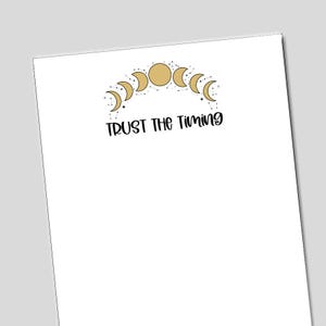 May include: A white rectangular sheet of paper featuring a graphic of gold moon phases arranged in an arc with small black star constellations. Below the moon graphic, the black text reads TRUST THE TIMING in a stylized, handwritten script font.