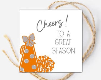 May include: Square white card with the text "Cheers! To a Great Season". The card features an orange megaphone with grey accents and a grey bow, along with an orange and grey pom-pom. A twine string is wrapped around the card.