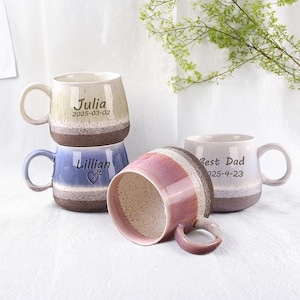 May include: Several ceramic mugs in shades of beige, blue, and pink, with brown bases. Each mug features a handle and personalized text, including names and dates. One mug is lying on its side, revealing its interior.