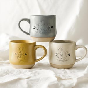 May include: Three ceramic mugs in grey, yellow, and cream. Each mug features a line drawing of a dog and cat with a heart and paw print. The mugs have a two-tone design with a light-coloured base and a curved handle.