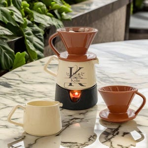 May include: A cream and brown ceramic coffee brewing set, including a pour-over cone, a carafe with a "Kylie" design, and a warming base. A small cream creamer and a brown pour-over cup and saucer are also visible. The set is designed for brewing coffee.