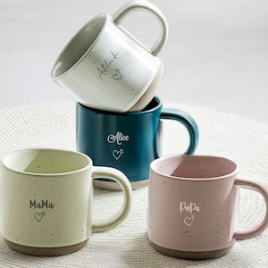 May include: Four ceramic mugs in different colours with names on them. The mugs are white, teal, light green, and pink. The names on the mugs are "Alice", "Mama", "Papa", and "Atlaude".