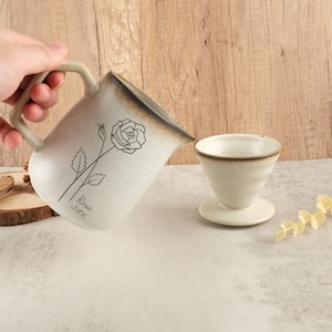 May include: White ceramic jug with a brown glaze and a black line drawing of a rose with the text "Rose June" on the side. The jug is being poured into a white ceramic pour-over coffee filter.