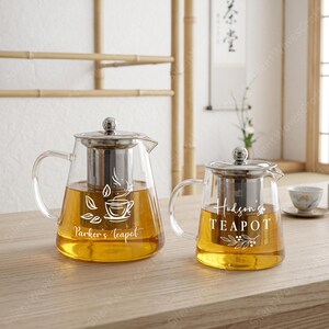 May include: Two clear glass teapots with stainless steel infusers and lids. The left teapot features a white graphic of a teacup with leaves and the text Parker's teapot. The right teapot displays the text Hudson's TEAPOT with a leaf branch design.