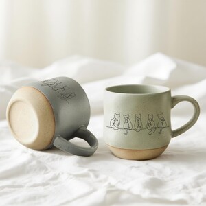 May include: Two ceramic mugs with a cat design. One mug is grey and the other is green. Each mug has a light brown base and a black line drawing of cats with names. The mugs are on a white surface.