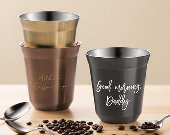 May include: Three stainless steel coffee cups in bronze, gold, and black. The bronze cup has the text "Arthur's Coffee Cup" in gold script. The black cup has the text "Good morning, Daddy" in white script. Coffee beans and spoons are also present.