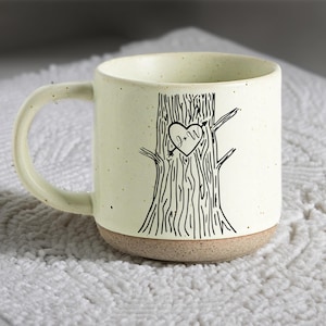 May include: Cream ceramic mug featuring a tree design with a heart containing the initials "J+W". The mug has a tan base and a curved handle. The mug is suitable for hot beverages.