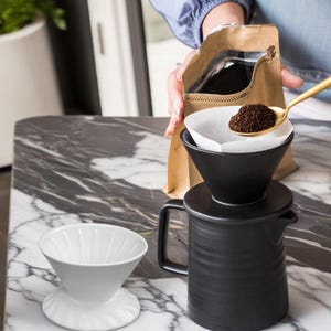 May include: A black ceramic pour-over coffee maker with a white ceramic filter and a white paper filter. The coffee maker is on a marble countertop. A person is pouring coffee grounds into the filter.