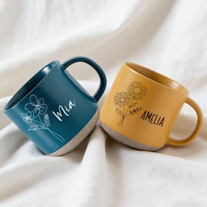 May include: Two ceramic mugs, one teal and one mustard yellow, against a white backdrop. The teal mug has a floral design and the name "Mia" in white. The yellow mug features a floral design and the name "Amelia" in black. Both mugs have a neutral-coloured base.