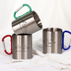 Könnte beinhalten: Drei Campingbecher aus Edelstahl mit Karabinergriffen in Grün, Rot und Blau. Jeder Becher zeigt ein Berg- und Baumdesign mit personalisierten Namen: Charlotte & Lucas, Elijah & Olivia und William.