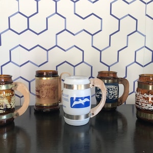 May include: Five brown glass mugs with wooden handles. Each mug has a different design featuring a location or landmark. The mugs are on a black surface.