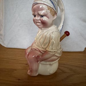 Puede incluir: Figura de cerámica vintage de un niño con gorro de dormir y camisón, sentado en un orinal. La figura está pintada con detalles en rosa, blanco y azul, y presenta un asa con un pomo rojo.