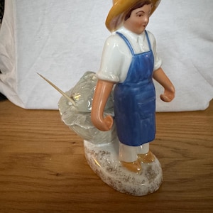 Pode incluir: Estatueta de cerâmica de uma pessoa com avental azul e chapéu de palha, ao lado de uma couve com um palito. A estatueta é pintada com detalhes realistas e assenta numa base com aspeto texturizado e envelhecido.
