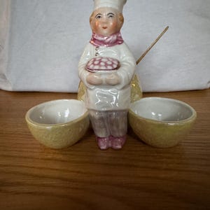 Peut inclure: Figurine de chef en céramique tenant une tarte, avec deux petits bols jaunes. Le chef porte un uniforme blanc, une écharpe rose et un grand chapeau blanc. La figurine est sur une surface en bois.