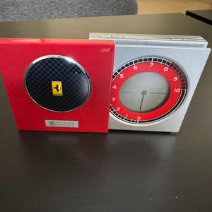 May include: A red and silver desk clock with a Ferrari logo. The clock has a round face with red accents and a black carbon fiber-style center. The Ferrari logo is a yellow prancing horse on a yellow background.