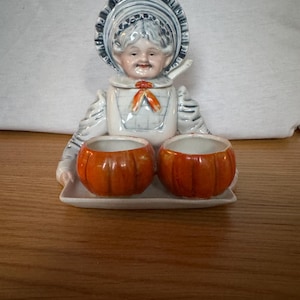 Peut inclure: Un service de table vintage en céramique. Il comprend un récipient en forme de femme avec un bonnet et deux bols en forme de citrouille. La figurine féminine est blanche avec des détails bleus et rouges. Les bols oranges reposent sur un plateau rectangulaire blanc.