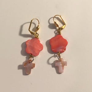 Peut inclure: Une paire de boucles d'oreilles pendantes avec des crochets dorés. Chaque boucle d'oreille comporte une perle en forme de fleur de couleur corail et une perle plus petite en forme de croix rose clair. Les boucles d'oreilles sont sur fond blanc.