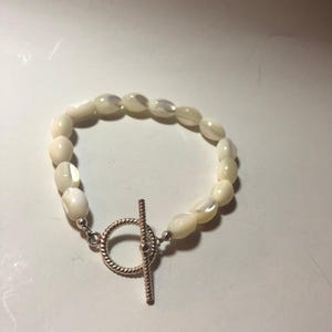 Peut inclure: Un bracelet composé de perles ovales blanc cassé. Il est doté d'un fermoir argenté avec un design circulaire et une fermeture à barre. Les perles ont un éclat nacré. Le bracelet est probablement destiné à la mode ou à la bijouterie.