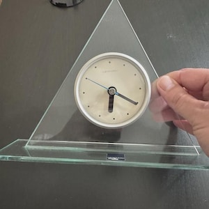 May include: A clear glass clock with a triangular shape and a round clock face. The clock face is off-white with black hour markers and black hands. The clock is labeled "LEONARDO".