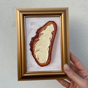 Puede incluir: Pintura al óleo enmarcada que representa una rebanada de pan con mantequilla. El pan es de color marrón oscuro y la mantequilla es blanca cremosa y espesa. El marco es dorado.