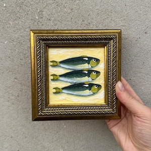 Può includere: Un piccolo dipinto quadrato in una cornice dorata ornata raffigurante tre pesci stilizzati con dorso verde scuro, pancia bianca e accenti gialli, disposti orizzontalmente su uno sfondo giallo pallido strutturato con pennellate visibili.