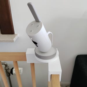 Può includere: Un baby monitor bianco con antenna grigia e staffa di montaggio bianca è attaccato a una sponda di culla in legno. Il monitor ha tre pulsanti e un piccolo obiettivo della fotocamera. Il nome del marchio è visibile sulla base.