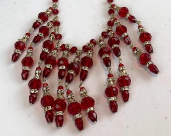 Puede incluir: Un collar de cuentas rojas con un diseño de borla. El collar está hecho de cuentas pequeñas y redondas y tiene una borla larga y colgante en la parte inferior.