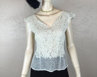 May include: A white lace top with a v-neckline and short sleeves. The top is fitted and has a scalloped hem. The model is wearing a black hat with a feather and a gold chain necklace.