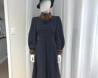 May include: A navy blue vintage dress with a faux fur collar and cuffs, displayed on a mannequin. The dress features a fitted bodice with bow details and long, puffed sleeves. A black hat completes the ensemble, offering a classic, elegant look.